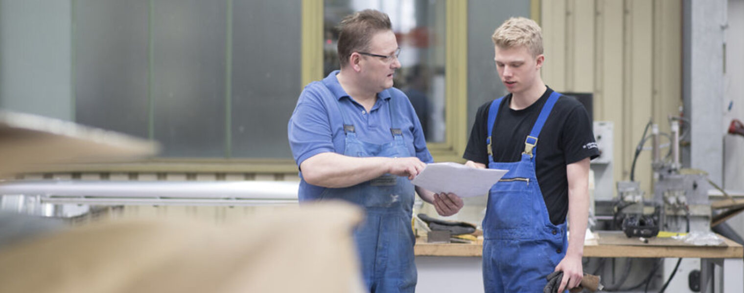 Ausbilder und Lehrling, Ausbilder zeigt dem Lehrling einen Plan, beide in Arbeitskleidung in einer Werkstatt