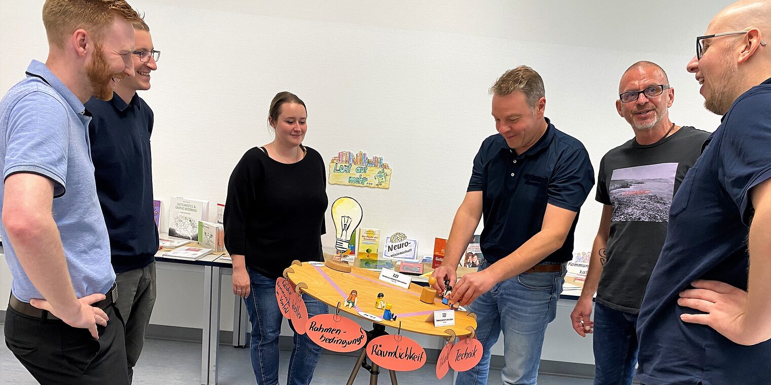 Die Kursteilnehmenden von Train the Trainer stehen um einen runden Tisch und tauschen sich über ein Thema aus.