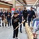 Hammer-Aktion beim Macher-Tag 2025 Besucher des Macher-Tags schlägt langen Nagel mit großem Hammer in Holz-Balken vor Publikum, das im Kreis steht.