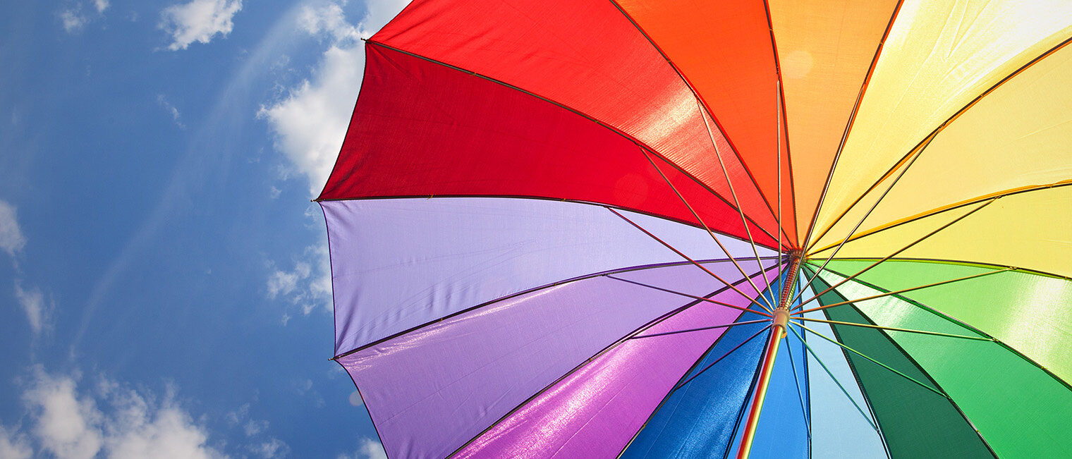 Bunter Sonnenschirm in Regenbogenfarben vor blauem Himmel mit Wolken