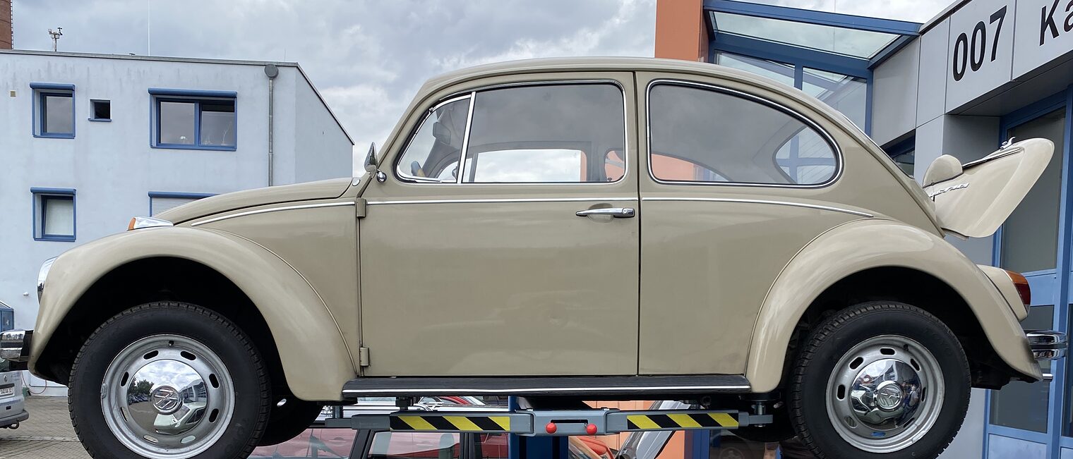VW Käfer Beiger VW Käfer steht auf Hebebühne vor Werkstatt, darunter grauer Volvo, zwei Personen arbeiten am geöffneten Motorraum.