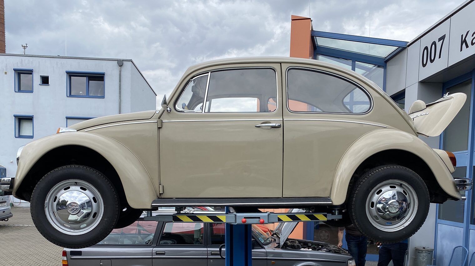 Beiger VW Käfer steht auf Hebebühne vor Werkstatt, darunter grauer Volvo, zwei Personen arbeiten am geöffneten Motorraum.