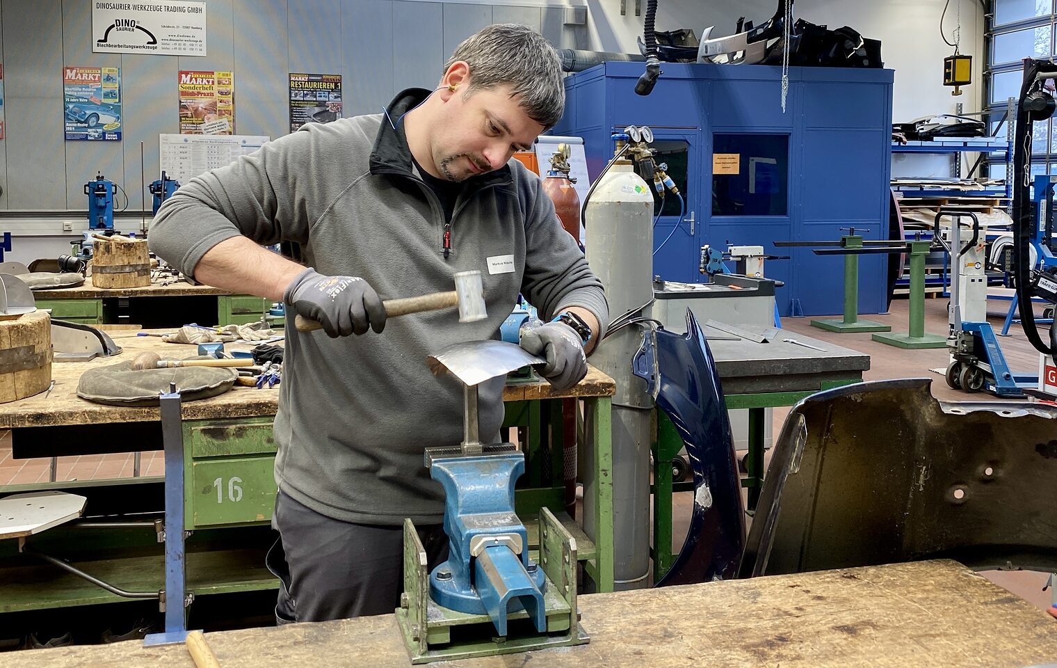 Blechbearbeitung Ein Mann bearbeitet ein eingespanntes Metallblech mit Hammer, trägt Handschuhe, im Hintergrund Werkzeuge und Gasflaschen.