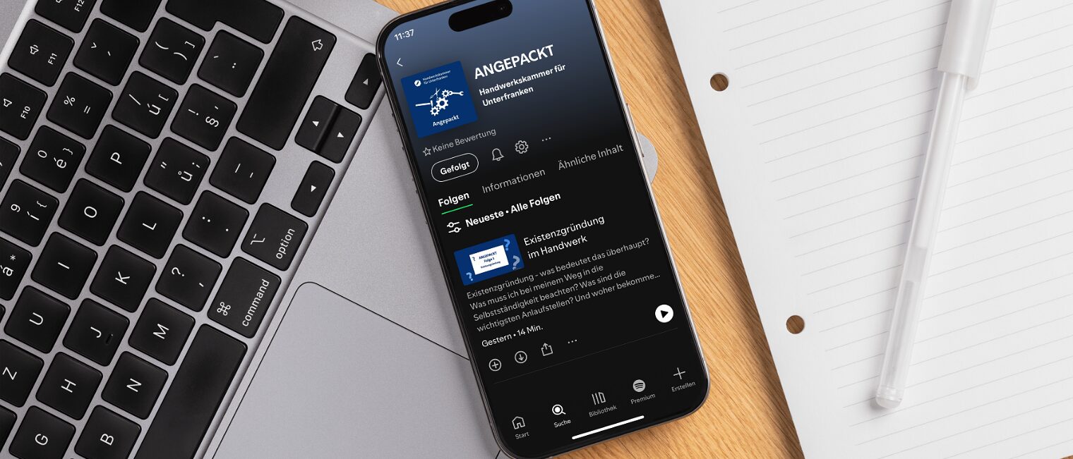 Smartphone mit Podcast "Angepackt" Smartphone mit Bildschirm, auf dem der Podcast "Angepackt" in Spotify geöffnet zu sehen ist, auf einem Schreibtisch.
