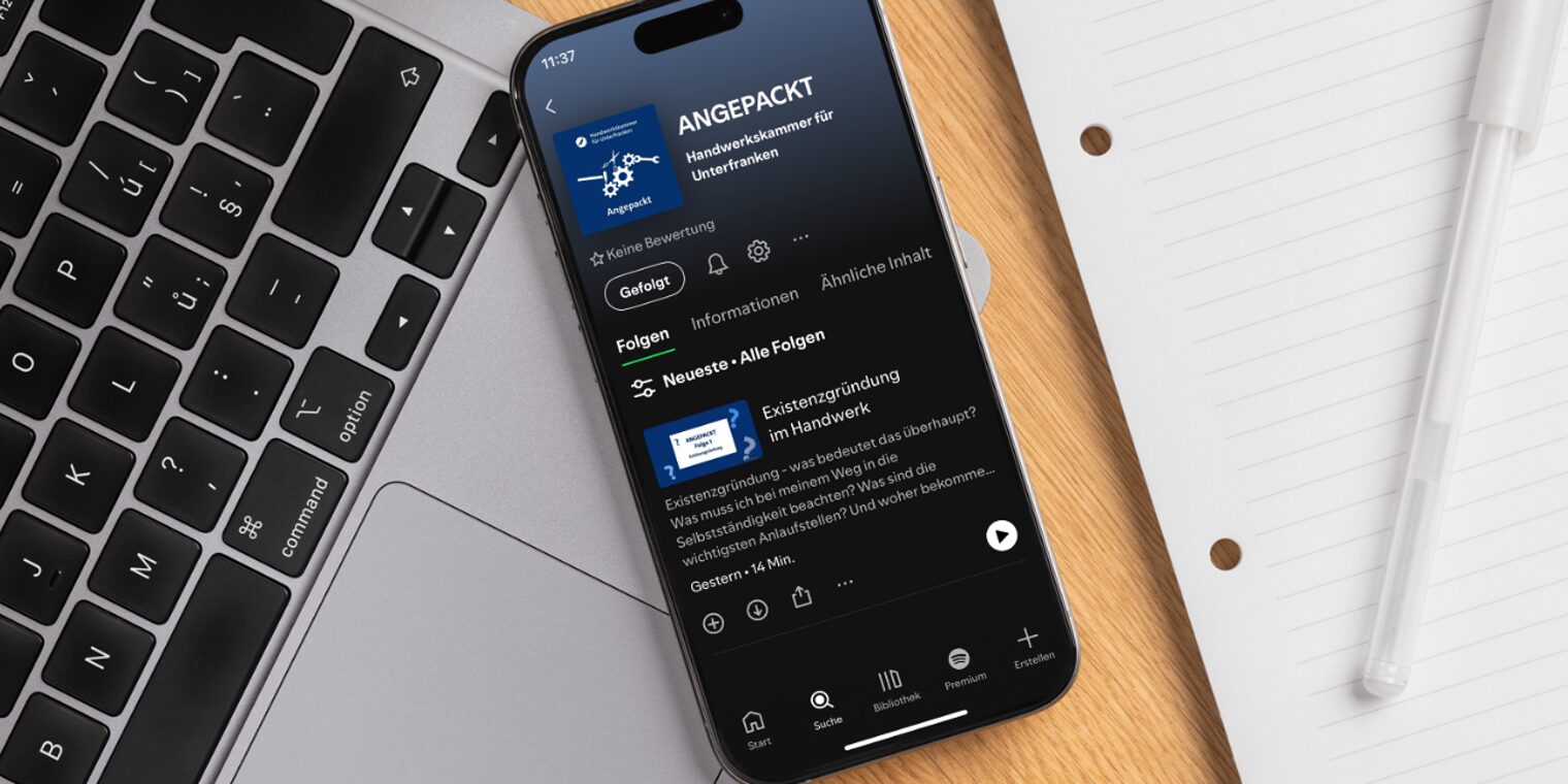 Smartphone mit Podcast "Angepackt" Smartphone mit Bildschirm, auf dem der Podcast "Angepackt" in Spotify geöffnet zu sehen ist, auf einem Schreibtisch.
