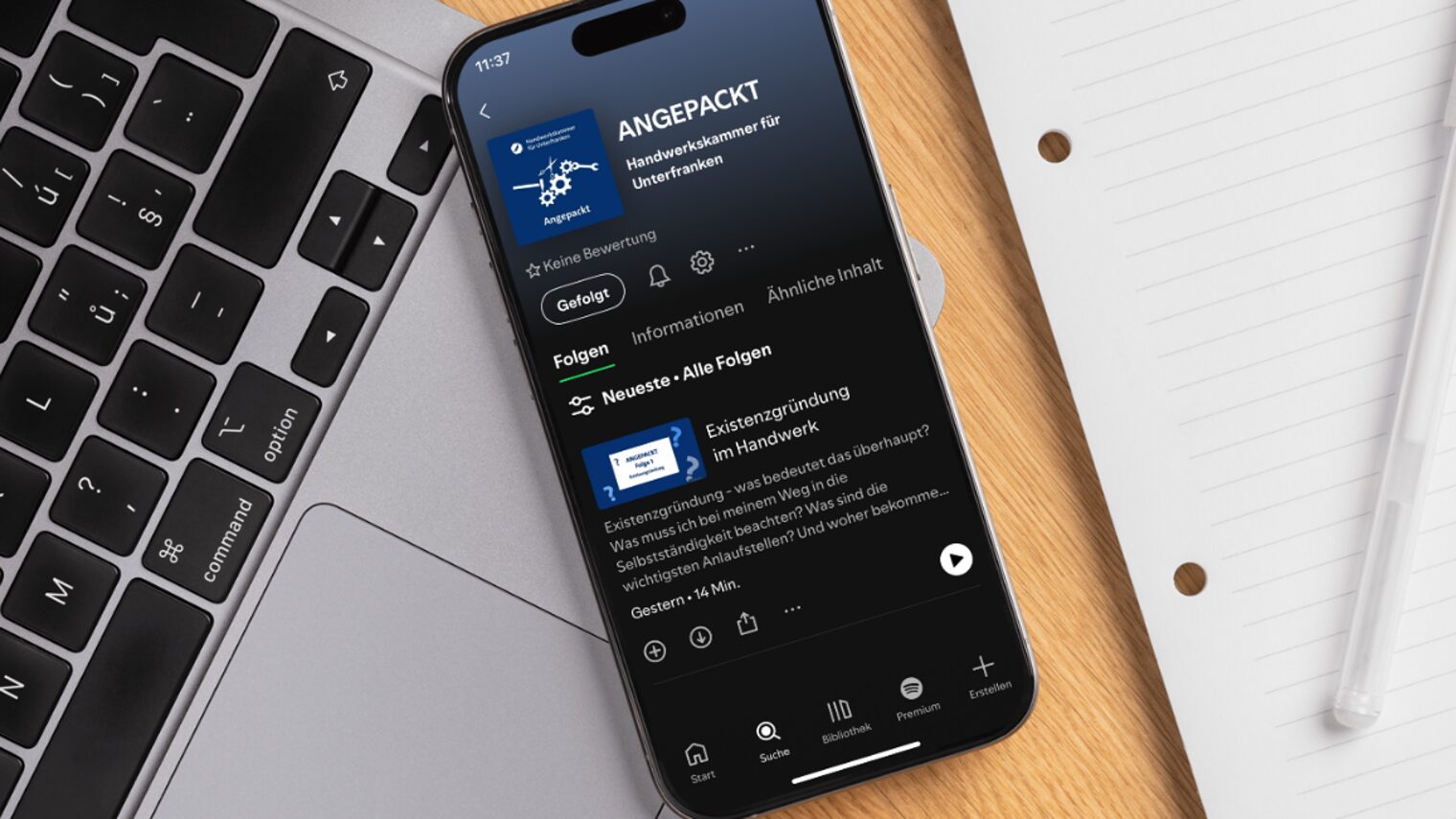 Smartphone mit Bildschirm, auf dem der Podcast "Angepackt" in Spotify geöffnet zu sehen ist, auf einem Schreibtisch.