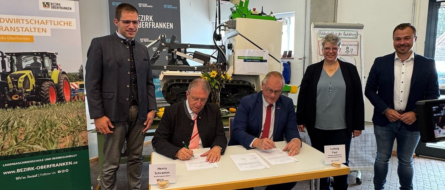 Fünf Personen sind auf dem Bild. Zwei sitzen am Tisch und unterschreiben einen Vertrag. Drei Personen stehendrumherum 