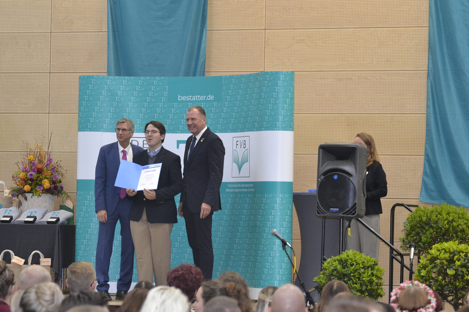 Drei Menschen sind zu sehen. Sie stehen auf einem Pult und einer bekommt eine Ehrung