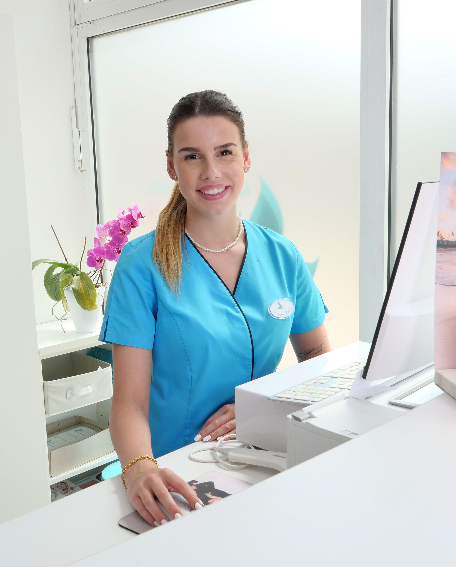 Frau in knallblauem T-Shirt am Tresen an einem Rechner, lächelt in die Kamera