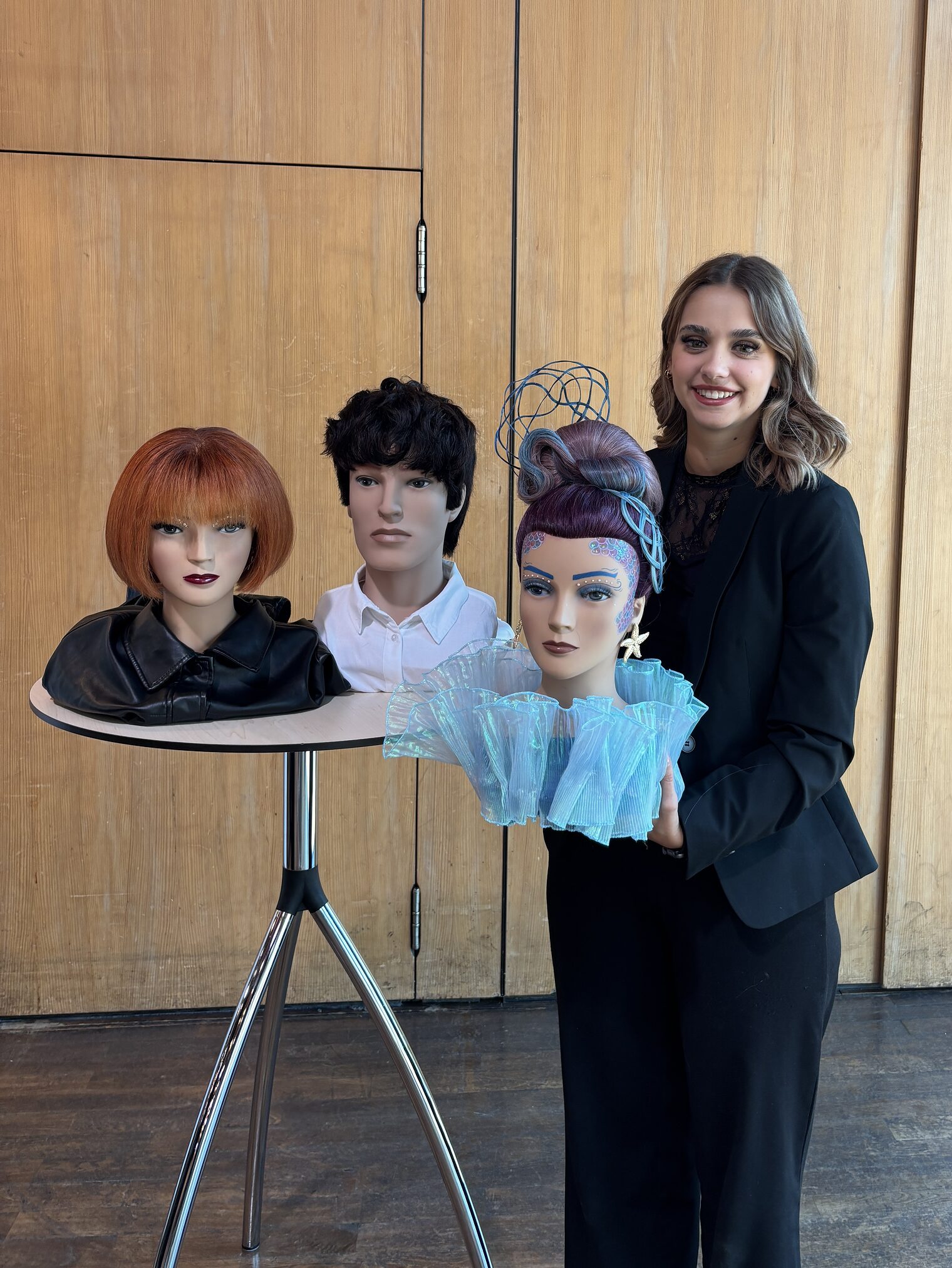 Junge Frau steht stolz an einem Stehtisch, darauf drei Frisierköpfe