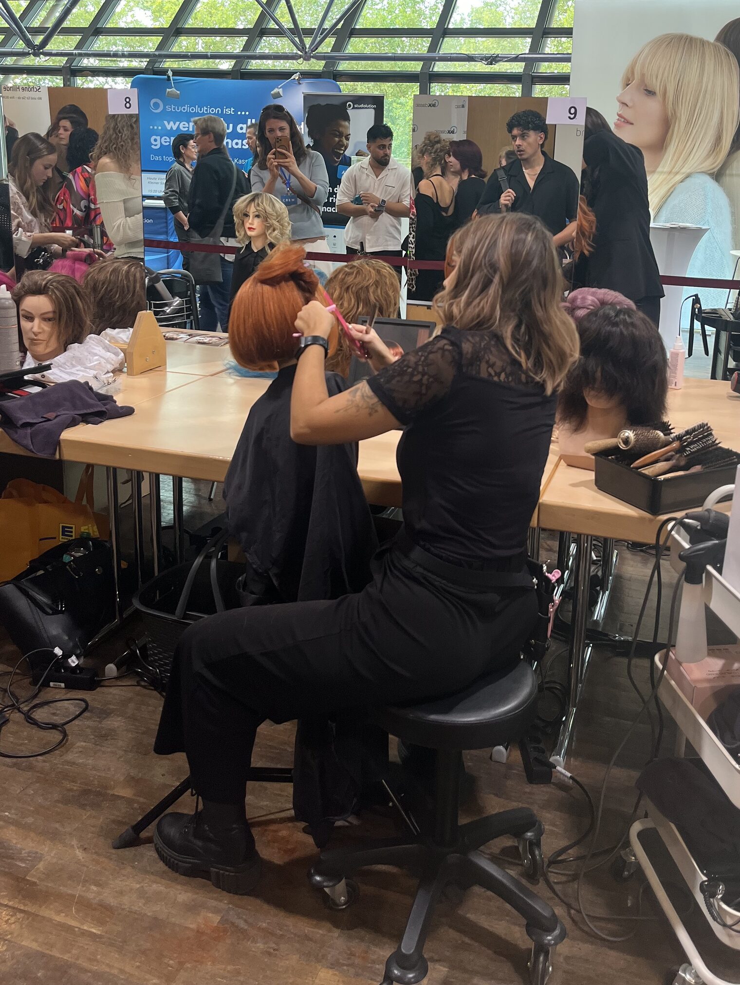 Junge Friseurin von hinten bei der Arbeit an einem Frisierkopf fotografiert. Im Hintergrund Wettbewerb mit Publikum und anderen Teilnehmerinnen.