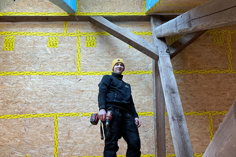 Zimmerer auf Baustelle unterm Dach, blickt nach unten in Kamera