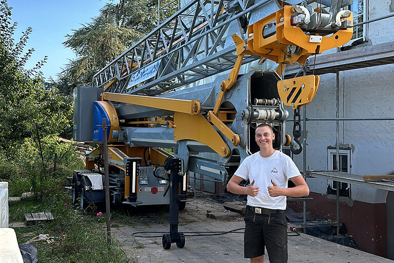 Zimmerer vor Baustelle mit Kran, zeigt Daumen hoch