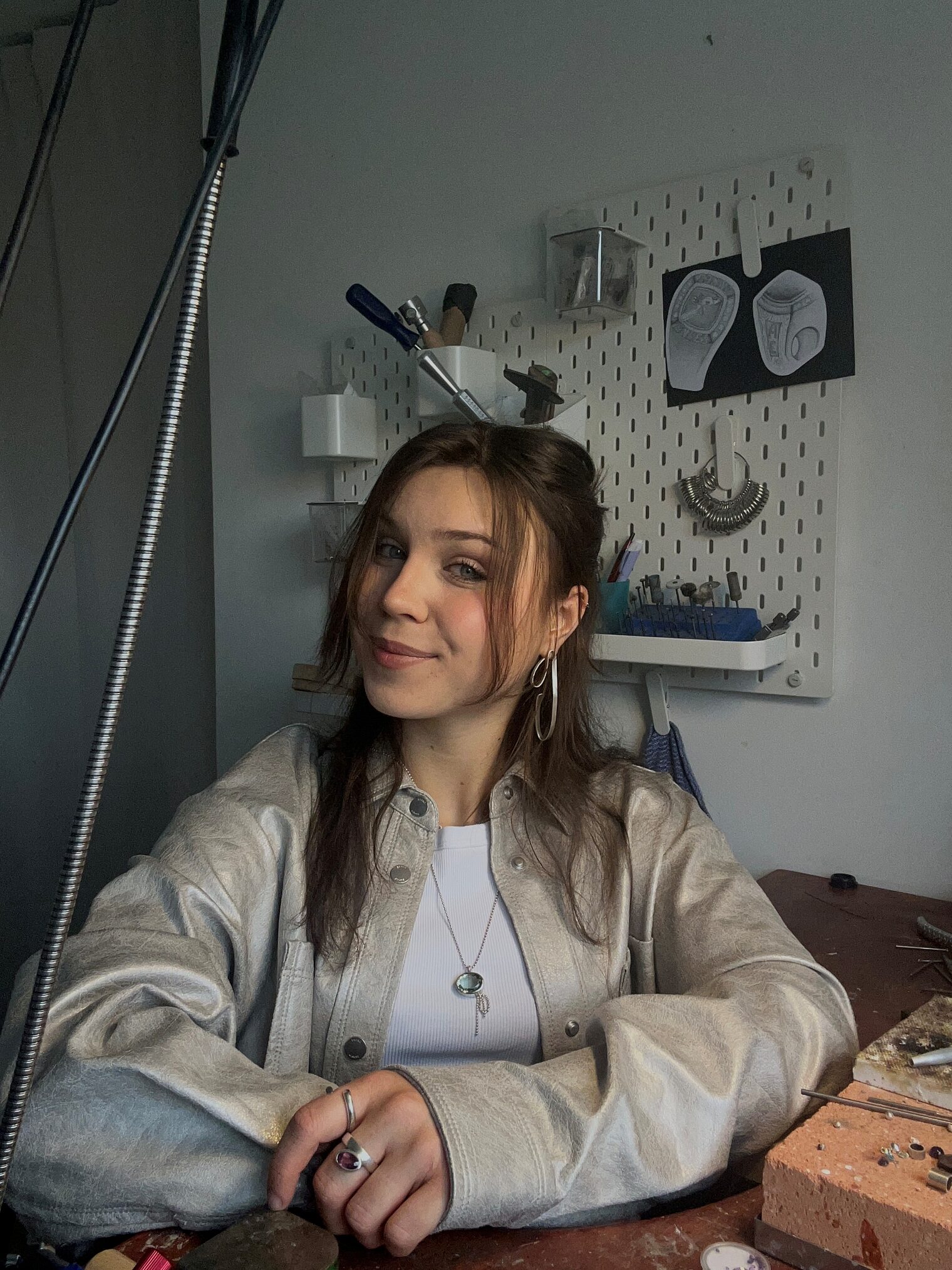 Junge Frau, braune Haare in heller Lederjacke, lächelt in die Kamera, Werkstatthintergrund Goldschmiede